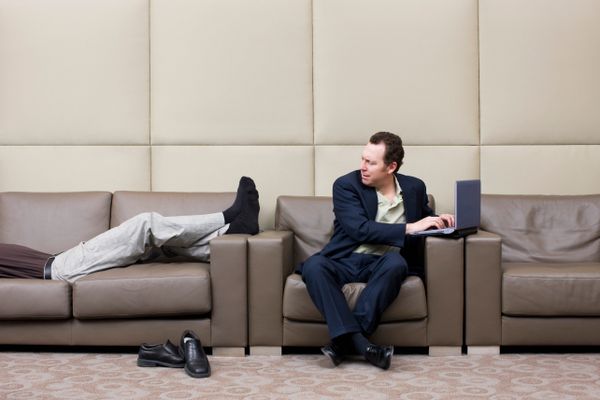 A businessman is annoyed by a man reclining on a sofa.