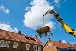 A crane lifts a prefabricated attic window into place on the roof of a prefab home.