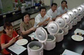 A group of official rice taste testers dip their chopsticks into five varieties of rice, prepared in the electronic rice cookers sitting in front of them.
