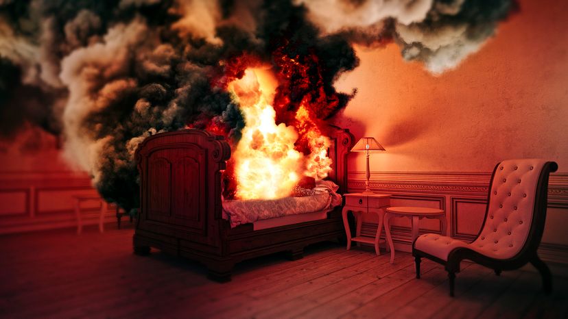 A bed, located in a bedroom with a chair, table, and bedside table, is on fire.