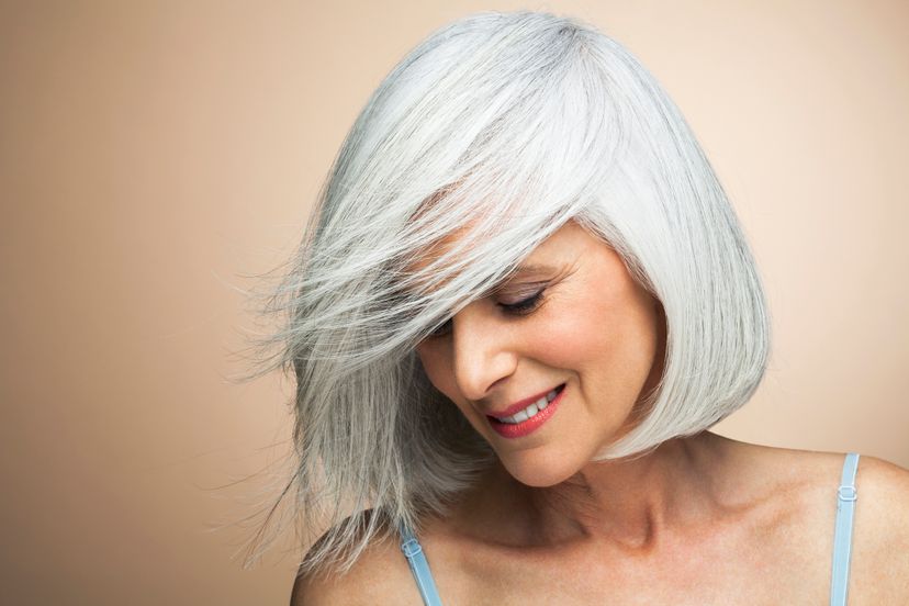 Mature woman with silvery, grey hair styled in a bob in front of beige background with wind blowing across her face looking down smiling.