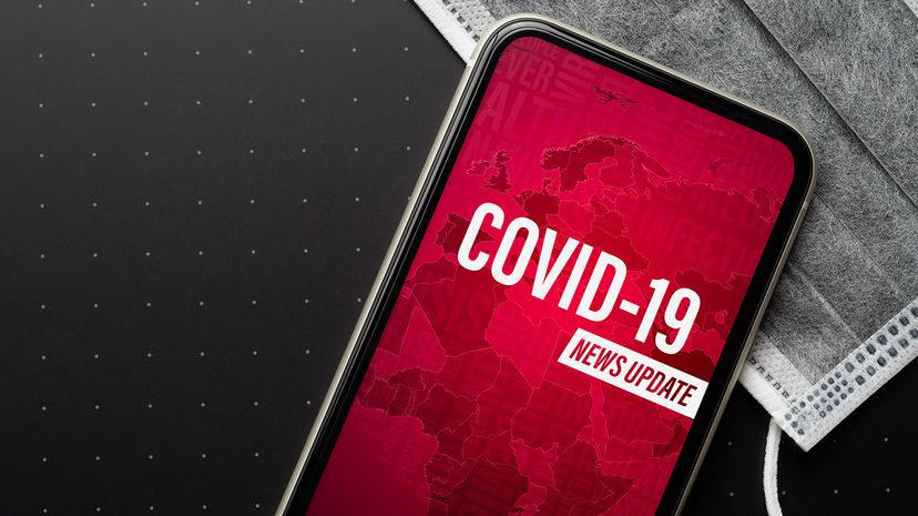 A phone on a grey surgical mask displays a red screen with white text about a COVID-19 news update.