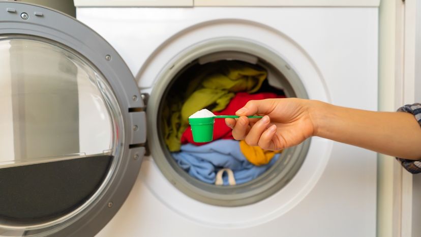 A woman's hand holds a cap with washing powder in front of a washing machine with clothes in it.  