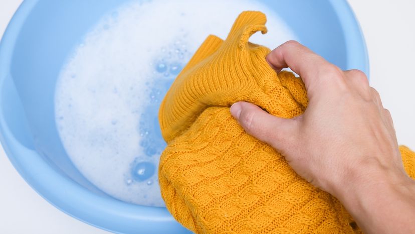 A hand adding a delicate mustard yellow cashmere sweater to a basin of soapy water.