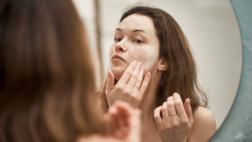 A woman washing her face with a washing foam while looking in the mirror.