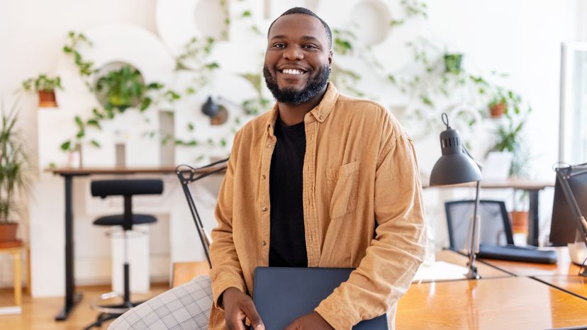 Portrait of a smiling businessman holding laptop leaning on a co-working desk in office.