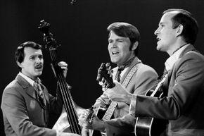 A black-and-white photo shows three musicians from the early 1960s singing.