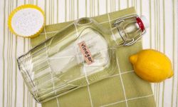 A bottle labelled "vinegar" laying on a green checkered cloth next to a lemon and baking soda.