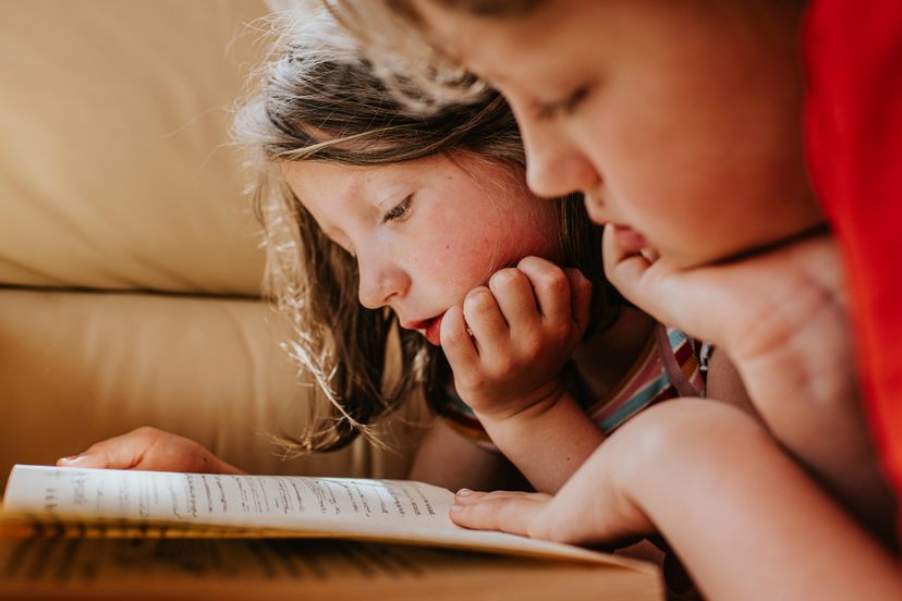 Two children share a book. They lie on their tummies, rest their heads on their hands and read the book together.