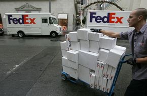 A Fed Ex employee wheels a dolly of parcels past two Fed Ex trucks.&nbsp;