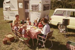 A family sits at a folding table eating a meal outside their station wagon and trailer, circa 1963.