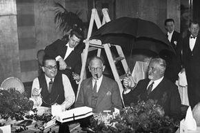 Three men sit at a table indoors, one of which is holding an open umbrella.