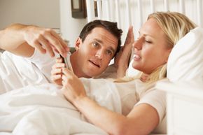 A man and woman are in bed; the man seems unhappy about the woman's use of a smartphone.