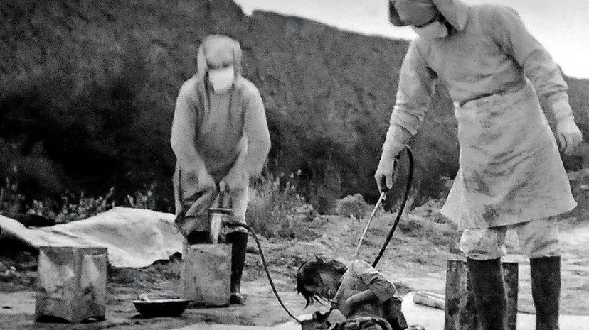 black and white photo of two people in PPE spraying bacteria on an unwilling person