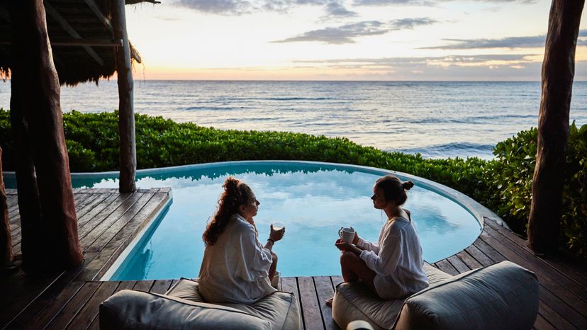 Female friends in discussion while sitting poolside at luxury suite of tropical resort at sunrise