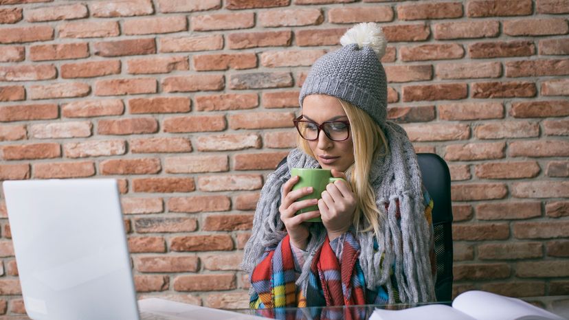 woman drinking hot tea and wearing hat and scarf at desk