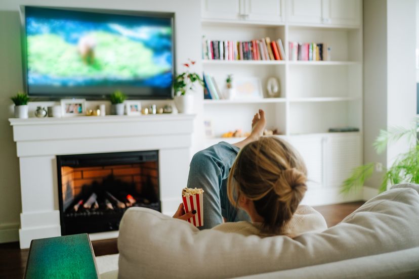 Blond woman with hair bun watching TV on a cozy sofa against fireplace indoors