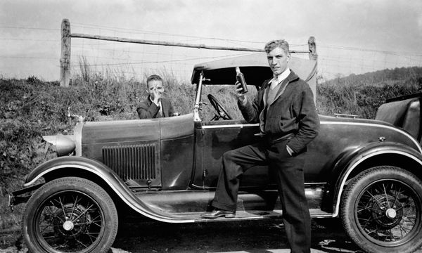 Two young men holding whiskey while standing at their car, c. 1930