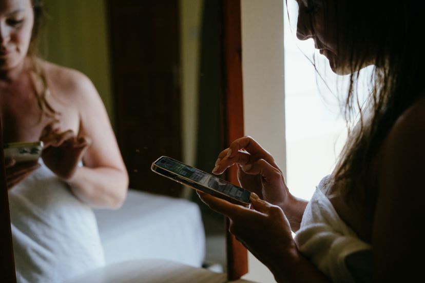 Woman in the hotel room with mobile phone texting and smiling, standing by the mirror with wet hair after a shower. Vacation in the tropical resort