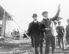 威尔伯·赖特（Wilbur Wright）使用风速计在飞行前检查风速。“border=