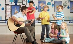 Male Teacher Playing Guitar With Pupils Having Music Lesson