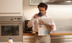 A man reading a newspaper in his kitchen while drinking from a mug.&nbsp;