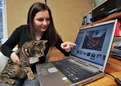 Ramona Markstein and her cat Fritz wearing a mini-camera around his neck sit in front of a laptop where pictures taken by Fritz are displayed.